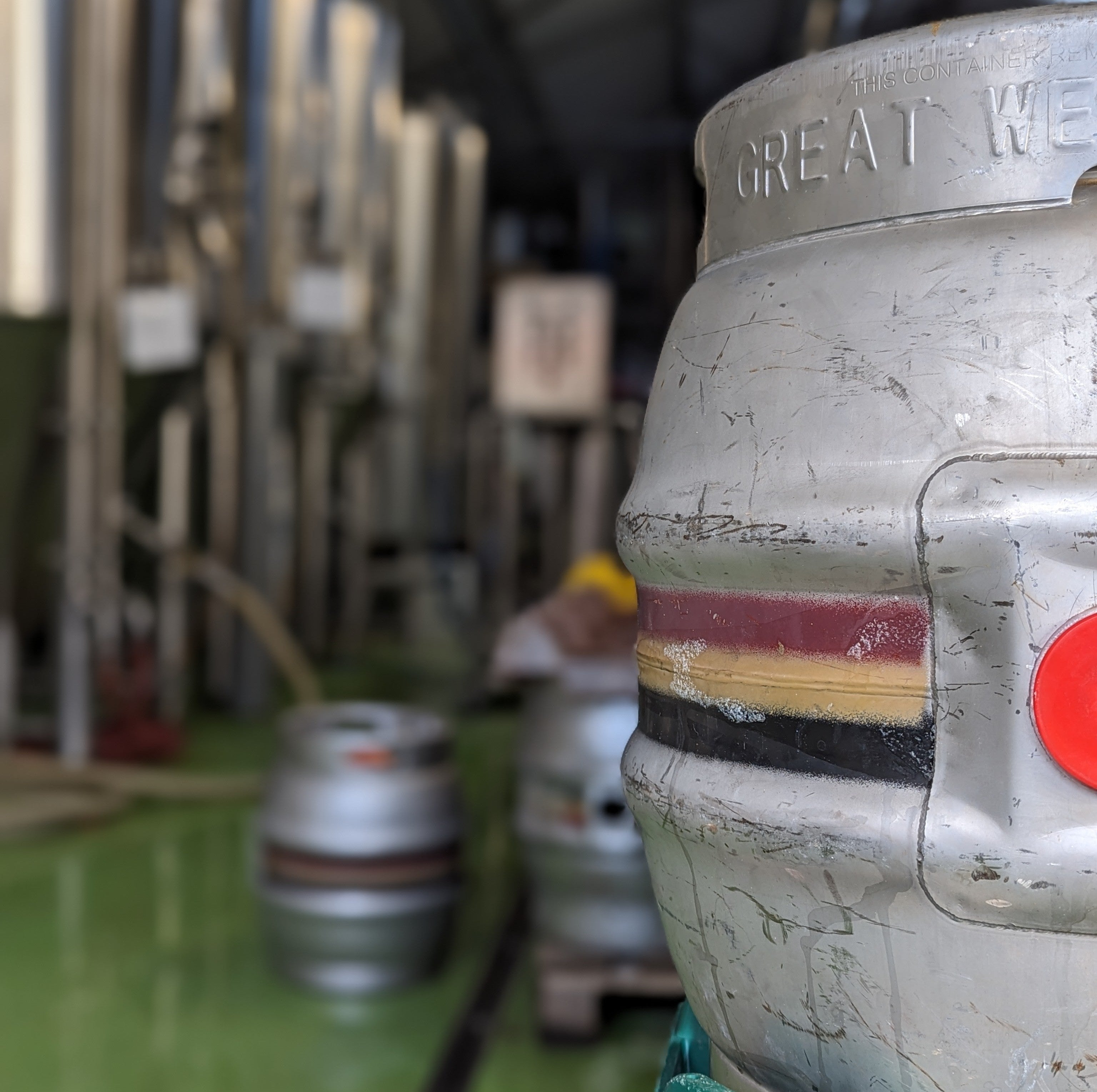A close-up of a metal beer cask with a branded red and yellow stripe design, with blurred figures and kegs in Hop Union Brewery in the background.