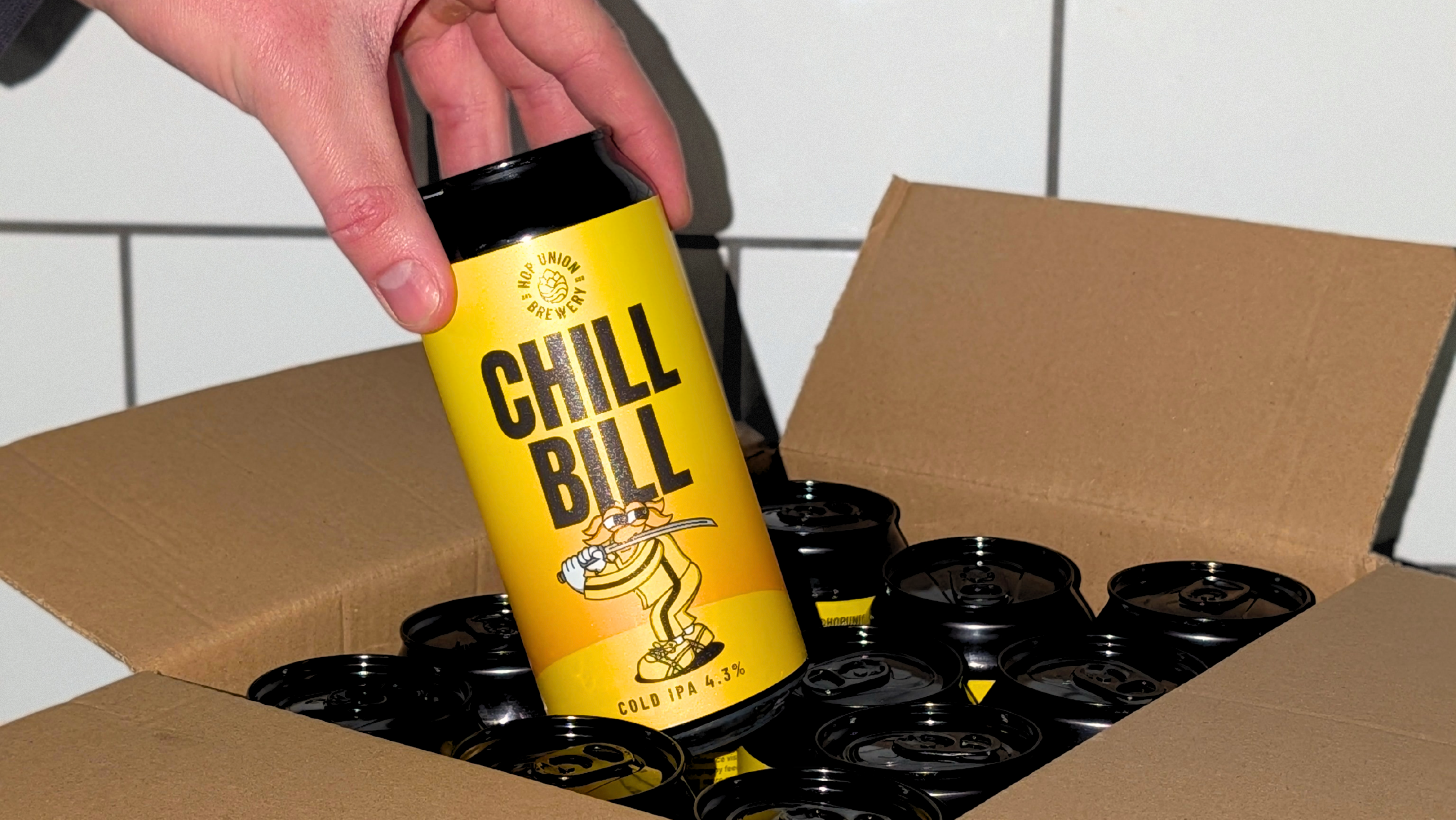 Person holding a 'Chill Bill Cold IPA' can above a cardboard box with more cans inside.
