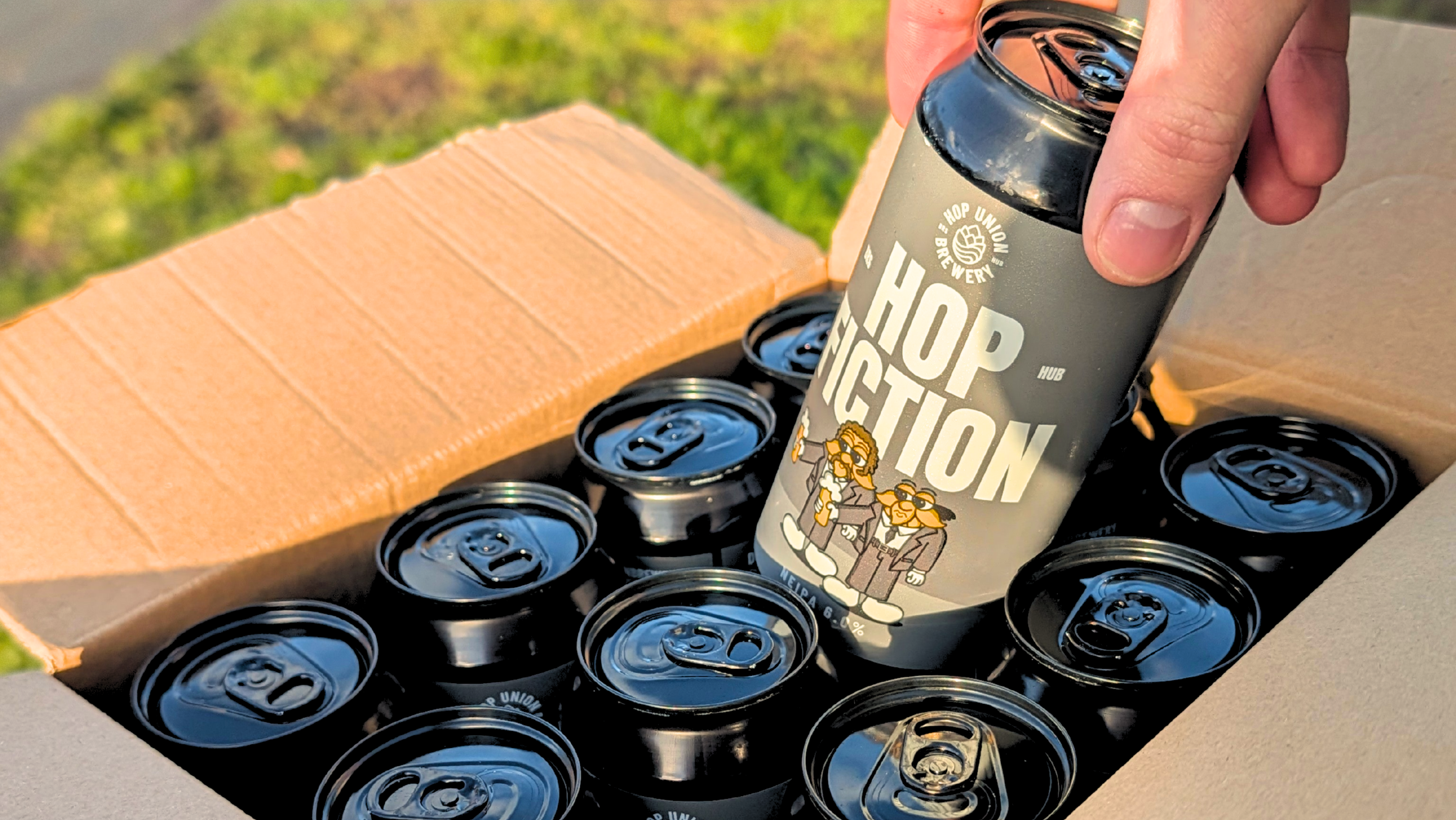 Person holding a can of Hop Fiction NEIPA over a box of cans.