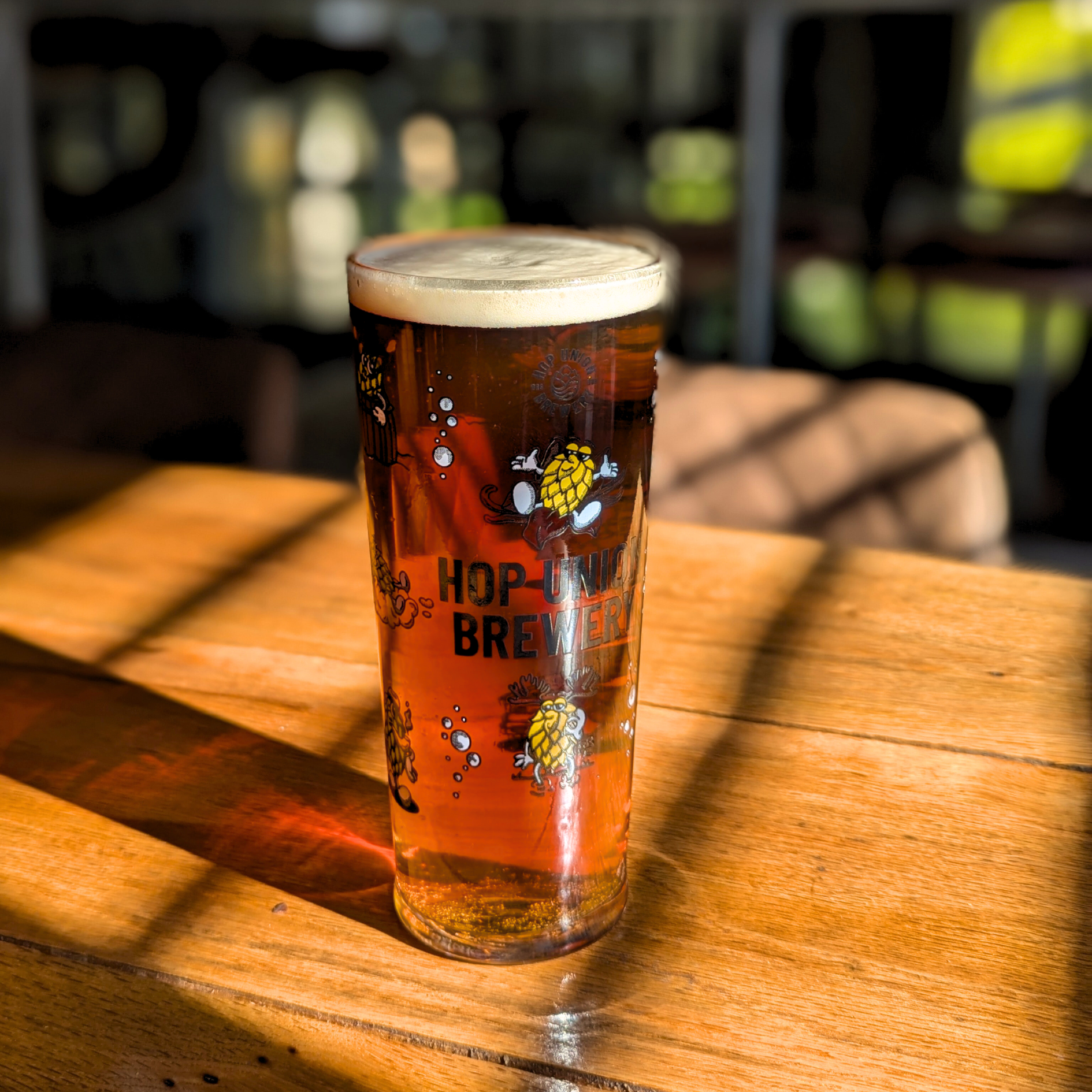 A Hop Union Brewery branded pint glass filled with Maiden Voyage Best Bitter on a wooden table in their Bristol taproom