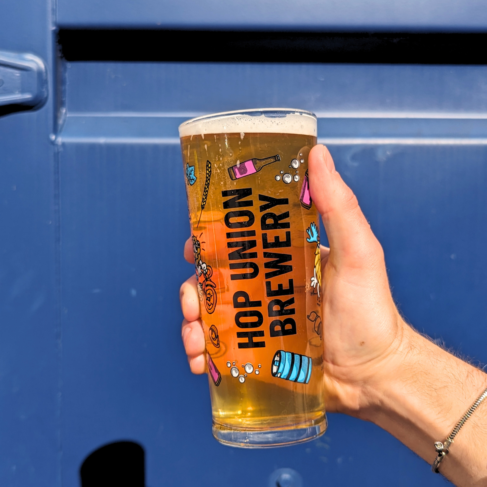 Hand holding up a Hop Union Brewery branded glass in front of a blue background