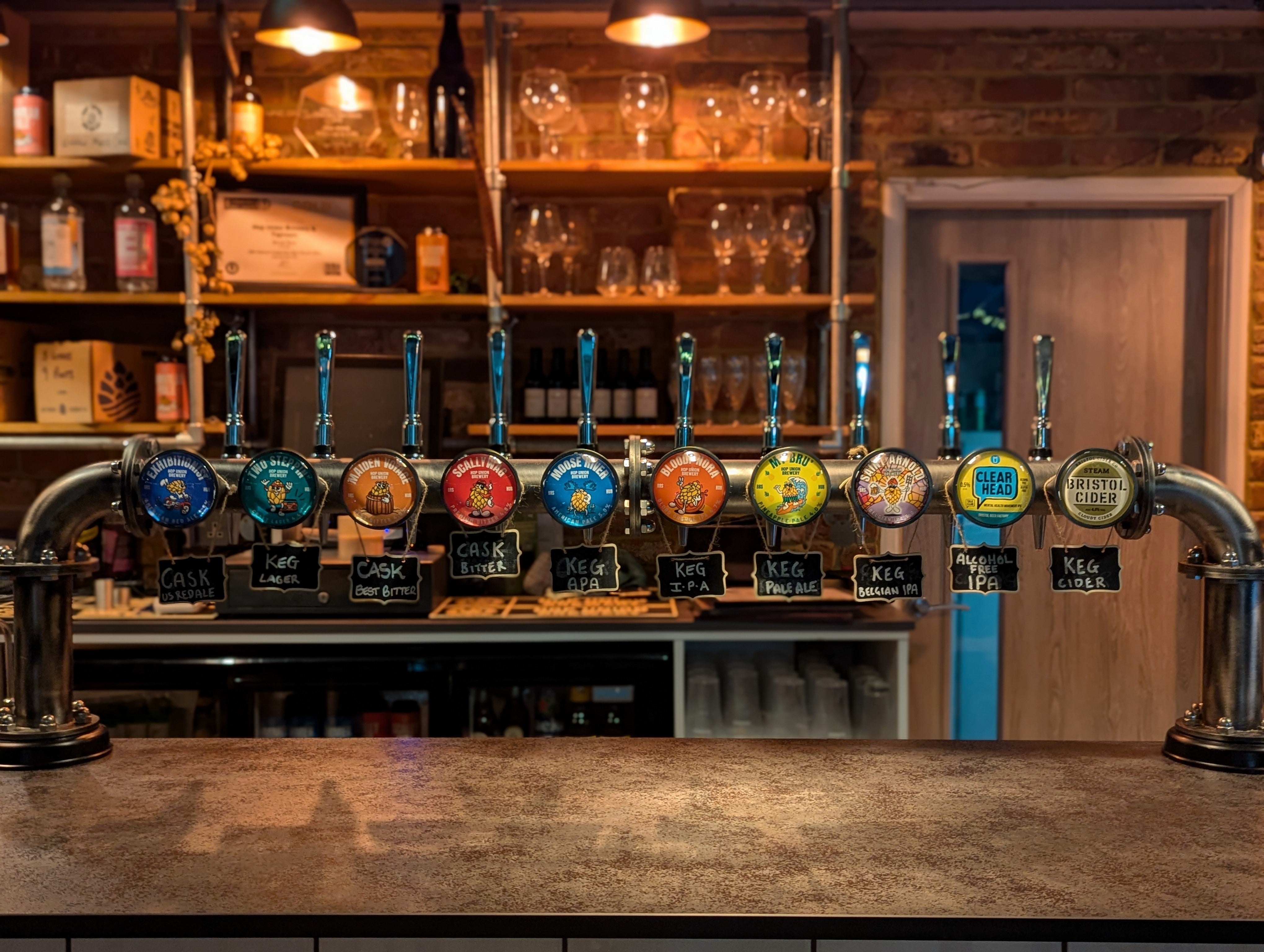 A bar font showing Hop Union Brewery beers on tap in the Bristol taproom with a blurred background of shelves and glasses