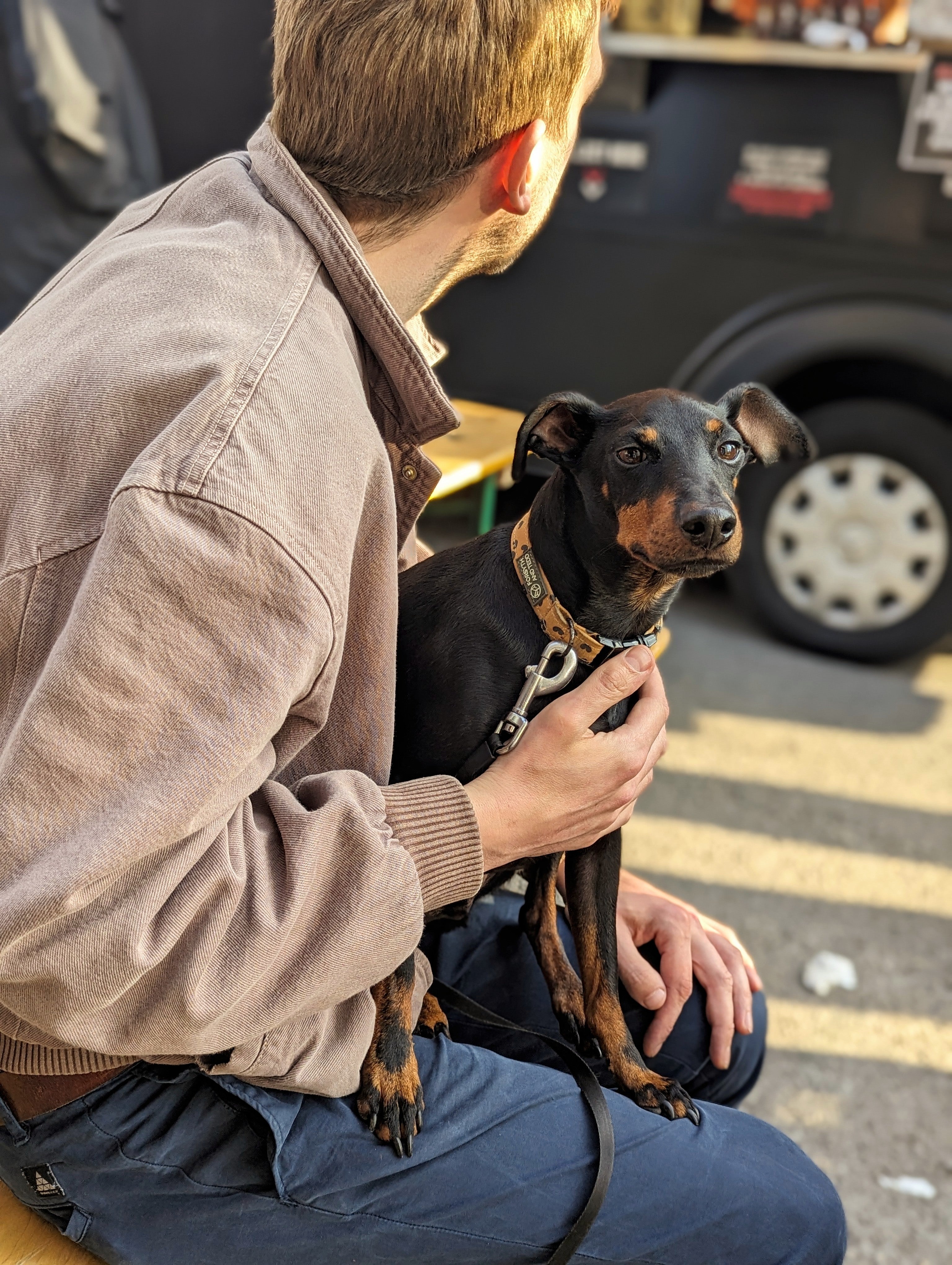 A photo of a man sat on a wooden bench outside the Hop Union Brewery taproom with a dog on his lap with a blurred background of a street food van